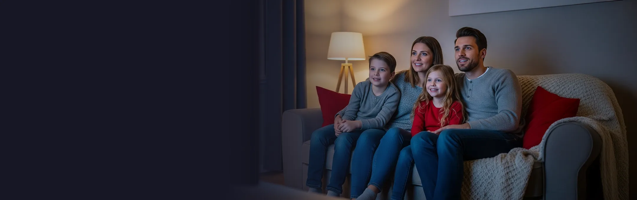 A happy family of four, including a mother, father, and two young children, sit smiling on a sofa in a dimly lit living room, watching television together.